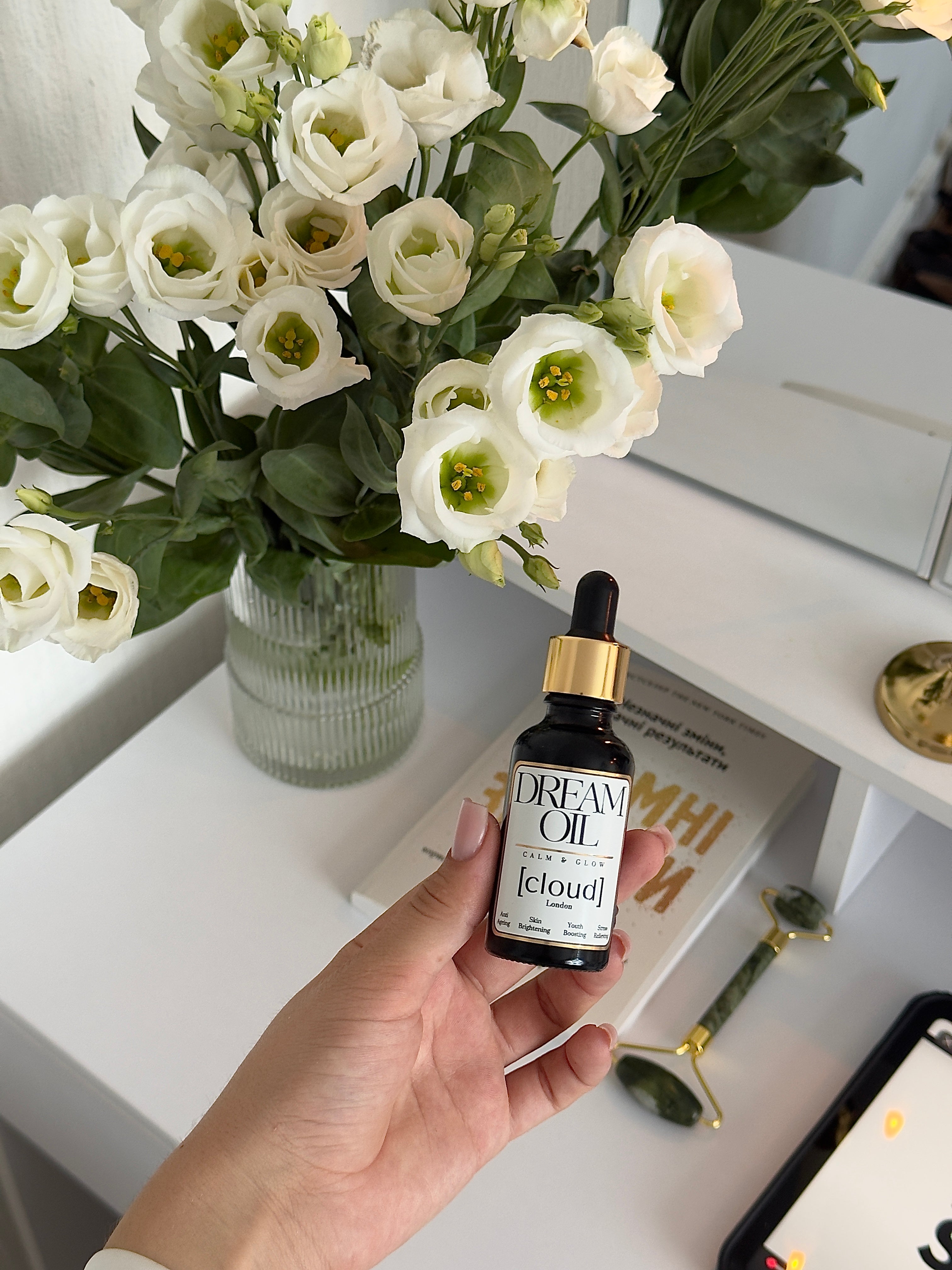 Hand holding a bottle of 'Dream Oil' with a vase of white flowers in the background.