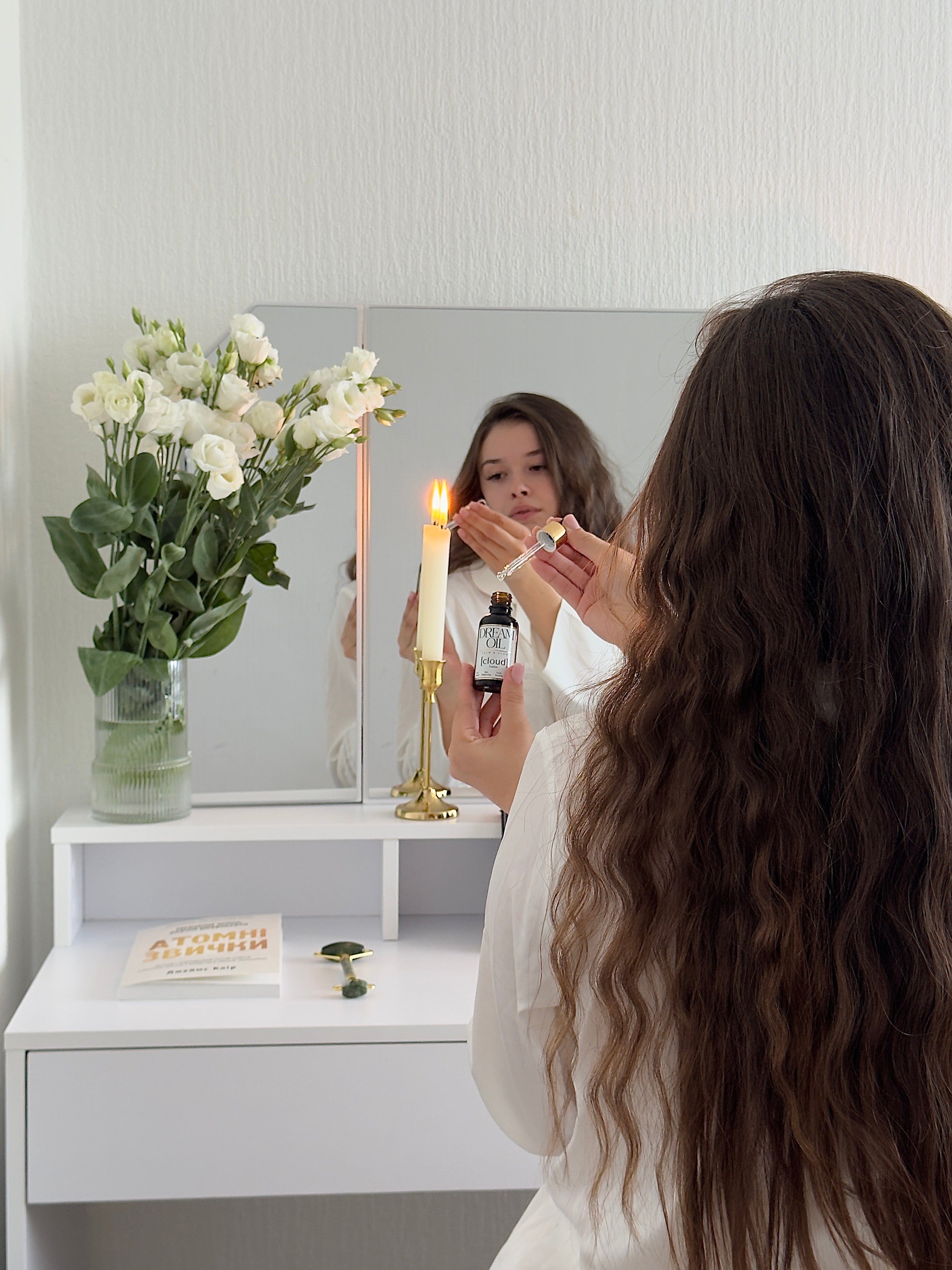 Woman applying Dream Oil in front of a mirror with a candle and flowers on a white dresser.